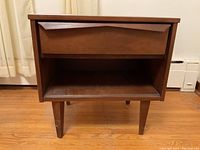 Front view of wooden cabinet showing one drawer and open shelf, positioned on hardwood floor.