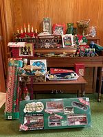 Photo showing full lot with various Christmas decorations on a piano and a table, including boxed Holiday Trains set, candle holder, ornaments, cards, and a tin train figurine.