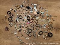 Top-down photo showing a large assortment of costume jewelry including many bracelets, earrings, necklaces, and rings spread on a wood floor.