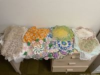 Photo of a collection of various crochet and lace doilies and table linens laid out on a table and dresser showing multiple colors and patterns.