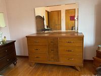 Full view of the wooden dresser with an attached mirror showing six wide drawers and three central smaller drawers or doors, placed against a pink wall on hardwood flooring.