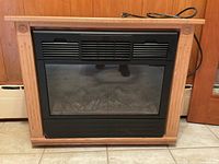 Frontal view of electric fireplace showing the wooden frame, black front vent panel, glass viewing window with faux logs inside, and power cord on tile floor.