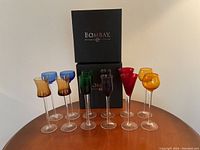 10 colorful cordial glasses of various colors arranged in front of 2 black boxes labeled Bombay.