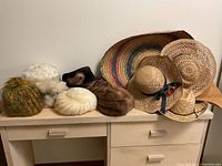 Photo showing all hats lined up on a table against a plain wall, including winter faux fur hats and knit hat on the left and straw and sombrero hats on the right.
