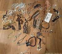 Photo showing wide assortment of costume jewelry items spread out on a wooden floor. Multiple necklaces and bracelets in various colors, materials, and styles visible.