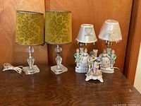 Two pairs of lamps and porcelain figurines shown grouped on a wooden surface against a wood backdrop. Clear square-based lamps have yellow-green floral fabric shades. Smaller footprint lamps have white decorative lampshades with figurine bases depicting colorful people. Additional separate porcelain figurines are present.