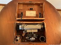 Wooden box opened showing the cylindrical surveying instrument and accessories inside with wear and damage visible.