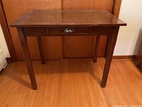 Front view of the wooden table showing the single drawer with metal cup handle and the four tapered legs on a hardwood floor against a wooden door background.