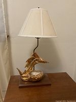 Full view of table lamp showing gold fish figurine base, curved metal stem and cream fabric shade on wooden platform