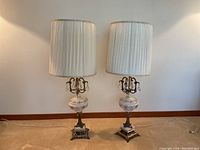 Pair of vintage table lamps standing side by side with pleated white fabric shades and ornate decorative bases involving porcelain and metal.
