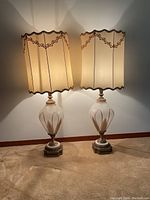 Pair of large vintage table lamps illuminated, showing scalloped cream fabric shades with decorative trim and ornate glass and metal bases.