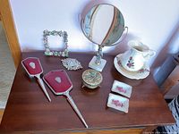 Photo showing the full vanity set on a wooden surface including hand-held mirror, brush, standing mirror, pitcher, basin, ornamental box, brooch, and decorative floral frame.