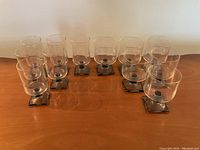 Set of 12 vintage stemmed glasses arranged in two rows on a wooden surface, clear glass bowls and square smoky gray bases visible.