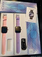 Two fitness watches in box packaging, one pink and one purple, with charging cable and manual included.