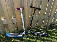 Two Razor scooters standing on grass against wooden fence, one white/blue electric and one black manual, showing overall condition and style.