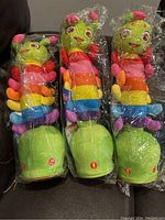 Three multicolored rocking caterpillar plush toys displayed side by side, wrapped in clear plastic.