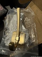 Single heavy brass-coloured wall hook with rectangular base plate and mounting hardware in plastic packaging placed inside a cardboard box.