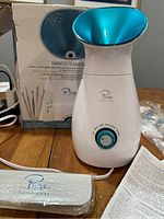 Front view of Nanosteamer facial steamer, showing teal steam outlet and dial control, box and instructions in background.