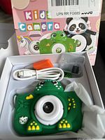 Front view of green frog-shaped kids camera in packaging showing included accessories and colorful box with animal graphics