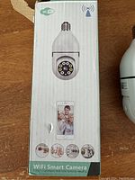 Front of box showing WiFi Smart Camera with camera lens, LED lights, and integration with a smartphone app for real-time remote viewing.