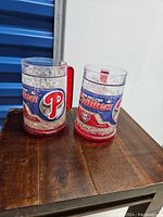 Two clear plastic freeze mugs with red handles and bases. Both mugs have Philadelphia Phillies 2010 graphics and logos. One mug filled with gel, the other one appears empty.