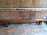 Photo of a long wooden mantel with carved decorative engravings, resting against a wooden wall. Shows surface scratches and wear.