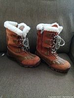 Front and side profile of Pajar women's boots in brown suede/leather with white faux fur at the top, lace-up front with black and white striped laces, and zipper on side.