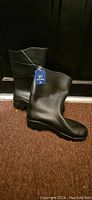Photo of a pair of black rubber rain boots with attached tag, standing upright against a dark background.