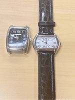 Two Bijoux Terner watches side by side on a wooden surface; one has a black leather embossed strap and white dial with black Roman numerals, the other has a black dial and silver case but no strap