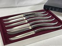 Six stainless steel steak knives neatly lined in custom burgundy fitting inside an open black box, showcasing curved handles and straight blades.