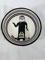 Top view of round clay dish showing black and cream paint with figure of person standing on caterpillar.