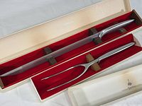Two-piece carving set with original boxes shown open, featuring a carving knife and carving fork in red velvet compartments, with cream exterior boxes, one box lid detached.