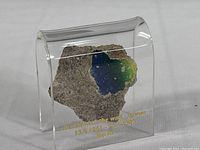 Front view showing the painted concrete fragment with blue and green paint enclosed in clear plastic display with gold date inscription