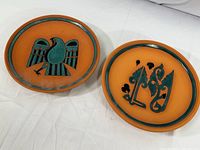 Pair of two 10 inch diameter terracotta decorative plates, one with a stylized bird motif and the other with abstract floral elements, both with black and turquoise painted accents.