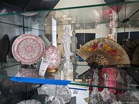 Glass shelf displaying an Ethan Allen decorative plate, matching egg figurine, white Asian figurine missing a hand, and a decorative fan.