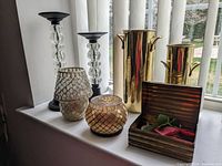 Overview of all items including two tall black and clear glass candle holders, two mosaic votive holders in front, two brass look handled metal vases behind, and a brass look trinket box open showing artificial greenery inside.