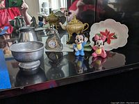 Various pewter-look items including a round bowl, pitcher, a horse-designed clock by Spoontiques, colorful Mickey and Minnie Mouse figurines, a small Lenox Winter Meadow floral bowl, and a mini teapot music box.