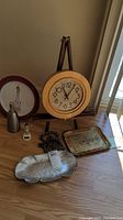 Wood round Seth Thomas clock on wooden easel, maroon Syracuse China plate with Space Needle illustration, painted wood tray featuring floral/landscape scene, metal floral tray, black Niagara Falls themed cast iron trivet, glass metal soap dispenser, small rooster kitchen timer on table.