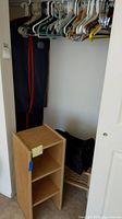 Photo showing wood-tone fiberboard 3-shelf cubby, hanging plastic and wooden hangers, black rolling bag, and plastic zip clothing bag hanging behind.