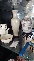 White ceramic vase, floral urn, candle holder, and porcelain bowl displayed on glass shelf