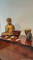 Wide view of all four items on display: large Buddha statue, lacquered salad bowl, decorative bowl with faux floral, and smaller metal Buddha head on stand.