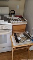 Photo showing assorted stainless steel utensils in white plastic trays on a kitchen counter next to a white stove, a metal teapot, and a small folding wooden table with additional kitchen tools.