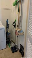 Photo showing Shark vacuum cleaner with multiple attachments along with Swiffer sweeper, Shark floor broom, Libman mop, mini step stool, Quickie mop, floor brush, and duster organized near a washer/dryer.