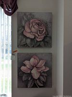 Two square floral prints on wood bases featuring pink roses with leaves, hanging on a wall with curtains beside them. Visible scratches on the surfaces.
