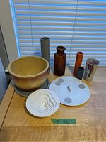 Photo shows nine assorted ceramic and glass items on a wooden surface near a window with blinds: a yellow-glazed bowl, multiple vases in grey, orange, and brown colors, a silver-toned cup with maroon speckles, a white ceramic decorative lid with gray circles and a handle, and a white multi-layered wave design ceramic piece.