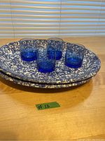 Two large blue and white ceramic platters with four blue lowball glasses arranged on top, displayed on wood surface in front of window blinds