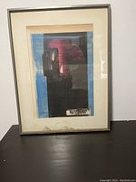Front view of framed abstract art showing mostly black and pink shapes with blue border inside a white matte and thin metallic frame. Artwork is off-centered inside the matte and frame.