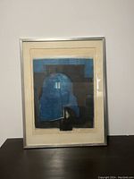 Full view of the framed abstract artwork showing blue and black geometric and organic shapes with a silver frame and off-white matte, displayed on dark surface against white wall.