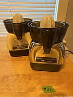 Two retro Proctor Silex automatic juicers side by side on table in front of window blinds. Juicers have plastic black tops with white reamers and mixed cream and metallic bodies with labels.