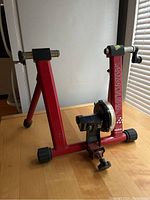 Red Minoura M80 stationary bike trainer on wooden table photographed from side angle showing full structure, roller mechanism and clamp knob.